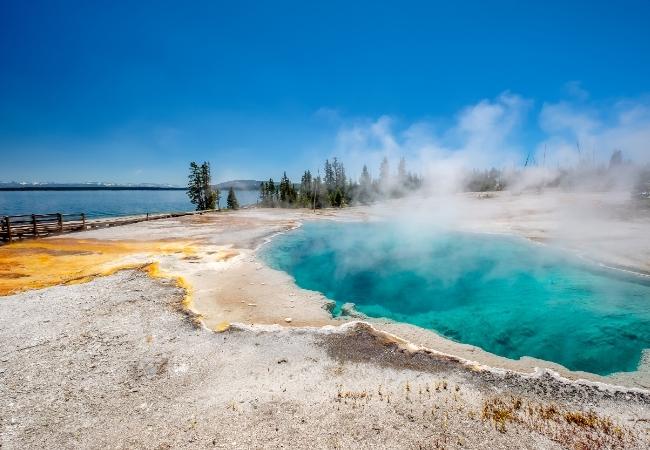 Eine türkisfarbene Thermalquelle dampft an einem felsigen Ufer am See unter tiefblauem Himmel.