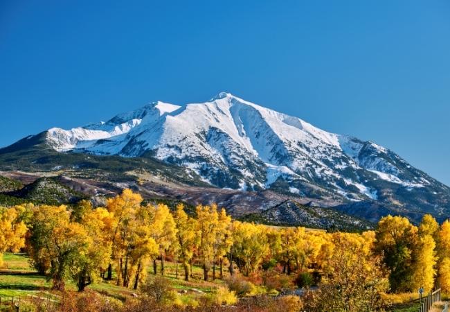 Schneebedeckte Rocky Mountains erheben sich über goldene Herbstbäume und offene Wiesen unter klarem blauem Himmel.