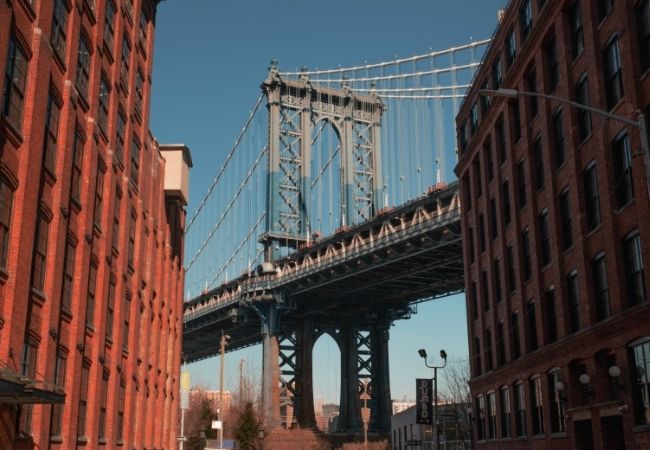 Zwischen roten Backsteinfassaden spannt sich die Manhattan Bridge über eine Straße in New York City unter klarem Himmel.