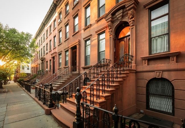 Reihenhäuser aus braunem Sandstein mit schmiedeeisernen Treppen und schmalem Gehweg in New York bei warmem Abendlicht.