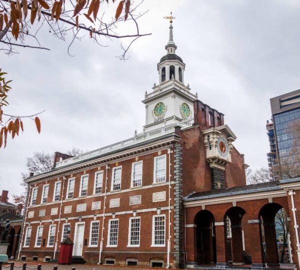 Vor einer hellen Wolkendecke steht ein historisches Backsteingebäude mit Uhrturm und Arkadengang in Philadelphia.