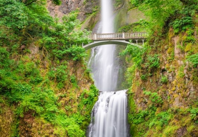 Ein hoher Wasserfall fällt durch einen moosigen Canyon, während eine schmale Bogenbrücke darüber führt.