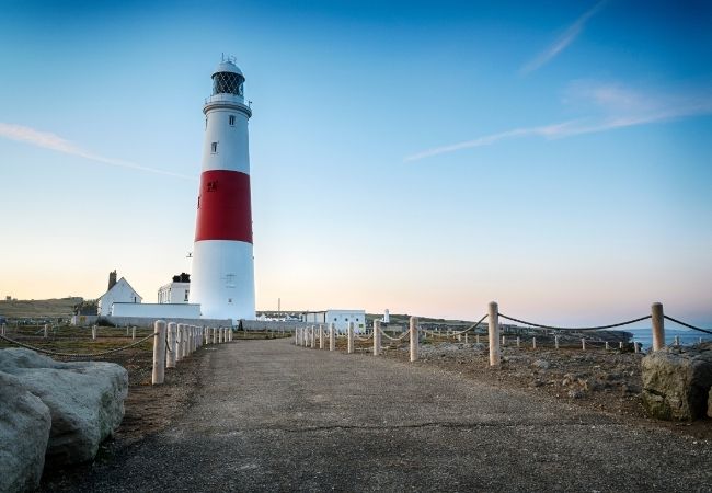 Ein rot-weißer Leuchtturm steht an einer felsigen Küste mit Weg und Seilpfosten unter klarem Abendhimmel.