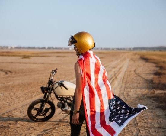 Ein Reisender mit goldenem Helm steht neben einem Motorrad in der Wüste, sein Cape in US-Farben weht im Wind.