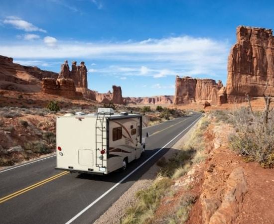 Ein Wohnmobil fährt auf der Route 66 durch eine weite Wüstenlandschaft mit hohen roten Felsformationen und blauem Himmel.