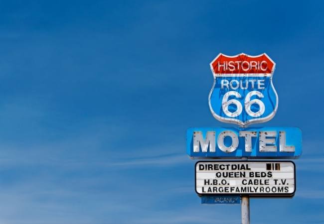 Vor blauem Himmel steht ein verwittertes Motelschild mit Historic Route 66, Motel und Direct Dial Queen Beds.