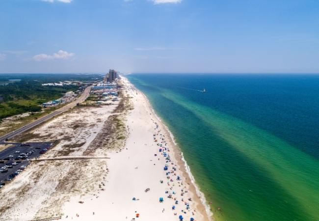 Eine lange Sandküste mit türkisblauem Wasser, Straße und Strandbesuchern zieht sich unter klarem Himmel entlang.