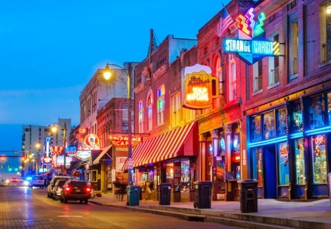 Eine neonbunte Straße in Texas zeigt Bars, alte Backsteinfassaden und zahlreiche Schilder im Abendlicht.