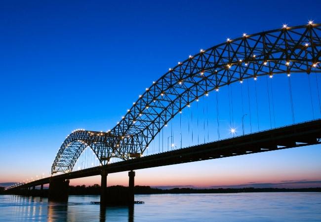 Eine beleuchtete Stahlbogenbrücke spannt sich bei Dämmerung über ruhiges Wasser unter klarem Abendhimmel.