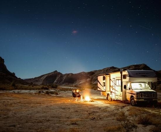 Ein beleuchtetes Wohnmobil steht nachts in einer weiten Wüstenlandschaft neben einem Lagerfeuer unter klarem Sternenhimmel.