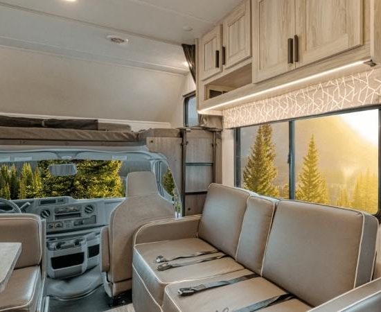 Der helle Wohnmobil-Innenraum zeigt eine Sitzgruppe mit beigen Polstern, Holzschränken und Blick auf die Fahrerkabine.