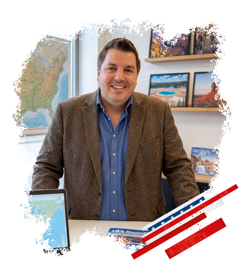 Im Büro des USA-Reisen-Experte steht Thomas vor Alaska-Karten, Reisefotos und einem Laptop mit Kartenansicht.
