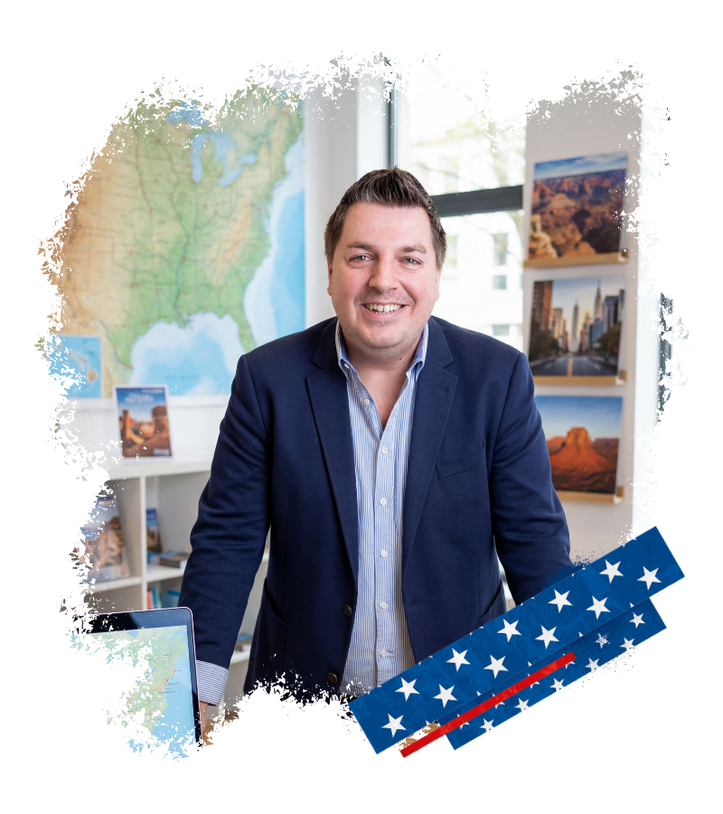 Thomas steht lächelnd im Büro von USA-Reisen Experte vor Alaska-Karte und USA-Reisezielen auf Posterwänden.