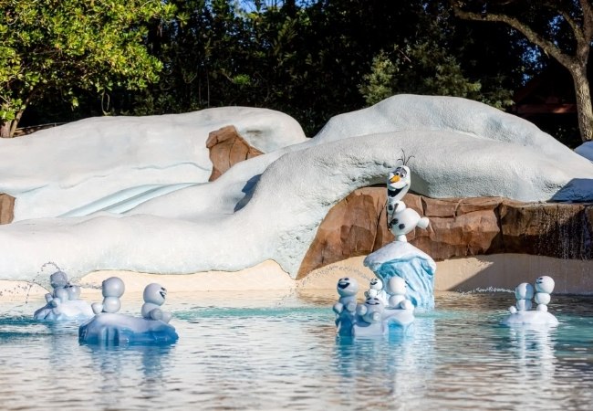 Im Disney Blizzard Beach ragen weiße Schneehügel, Eisskulpturen und eine Olaf-Figur aus dem türkisfarbenen Wasser unter sonnigem Himmel.