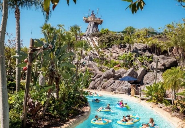 In einer tropischen Lagune von Disney's Typhoon Lagoon treiben Besucher auf Reifen zwischen Palmen, Felsen und einer wilden Rutsche unter blauem Himmel.