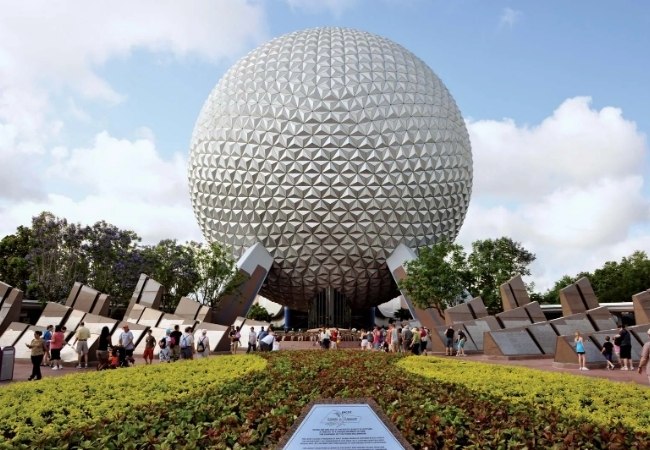 Vor dem silbernen Epcot-Kugelbau laufen Besucher über die Plaza unter blauem Himmel im Walt Disney World Resort in Orlando, Florida.