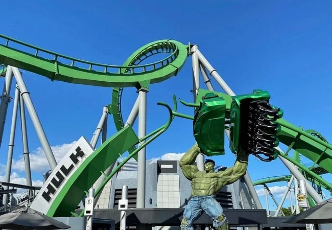 Vor tiefblauem Himmel ragt The Incredible Hulk Coaster in Islands of Adventure auf, daneben steht die grüne Hulk-Statue in Orlando.
