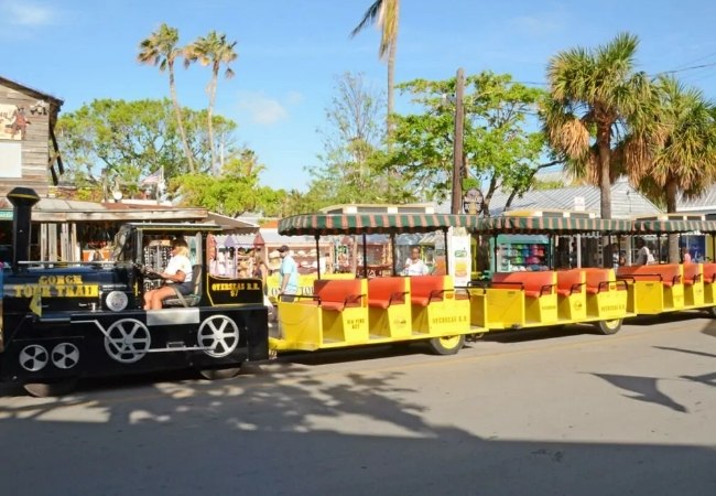 Ein bunter Old Town Trolley mit gelb-roten Waggons fährt durch die sonnige Straße von Key West, Florida, vorbei an Palmen und kleinen Shops.