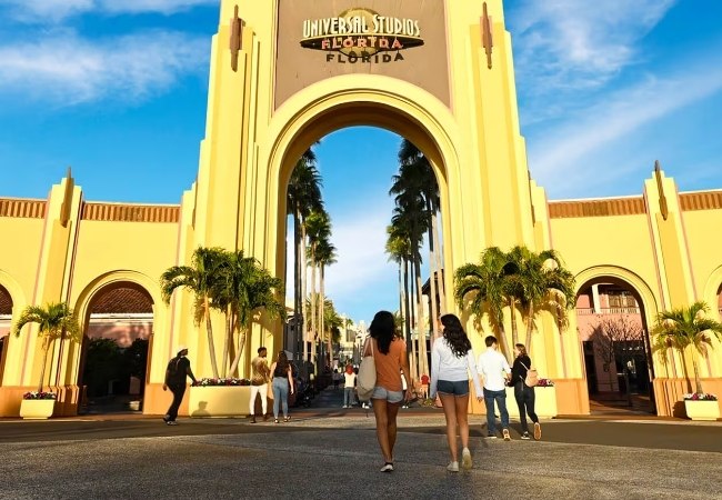 Im sonnigen Florida führt ein gelbes Tor von Universal Studios Florida zwischen hohen Palmen in den Park, während Besucher hindurchgehen.