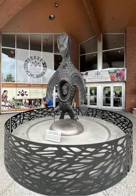 Vor dem Alaska Native Heritage Center steht eine dunkle Skulptur in einem runden, dekorativen Zaun vor dem Gebäudeeingang.
