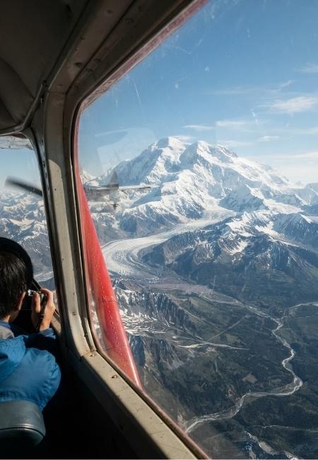 Aus dem Flugzeugfenster fällt der Blick auf den schneebedeckten Denali über einer weiten Gebirgslandschaft in Alaska.