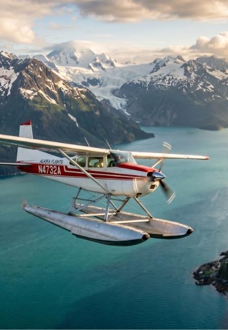 Ein rot-weißer Wasserflugzeug fliegt über türkisfarbenes Wasser vor verschneiten Bergen und einem Gletscher in Alaska.