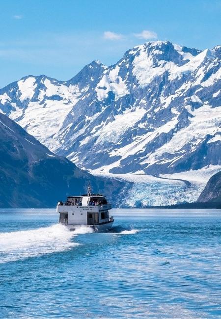 Ein Boot fährt durch türkisblaues Wasser vor einer großen Gletscherfront und schneebedeckten Bergen in Prince William Sound.