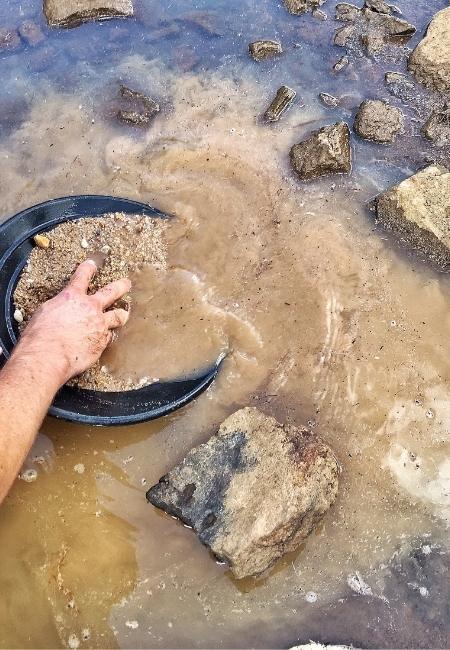 Eine Hand siebt mit einer schwarzen Pfanne Kies und Sand im trüben Flusswasser zwischen nassen Steinen.