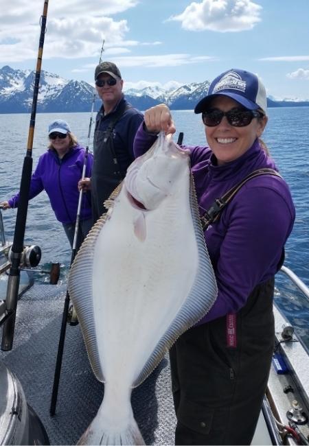 Drei Angler auf einem Boot in Alaska halten einen großen Heilbutt vor schneebedeckten Bergen und ruhigem Wasser.