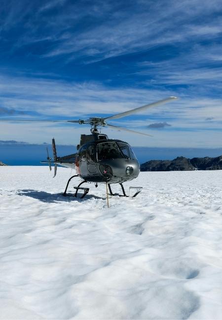 Ein Helikopter steht auf einem Schneefeld vor Meer und Bergen unter blauem Himmel bei einer Gletscherlandung in Alaska.