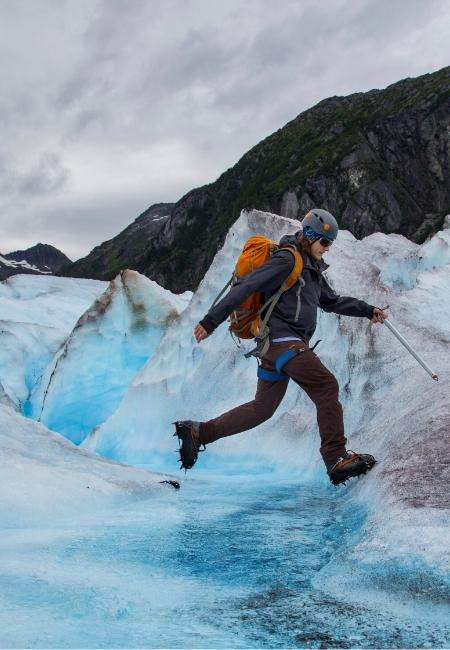 Eine Person mit Helm und orangefarbenem Rucksack springt über blaues Eis am Matanuska Glacier in Alaska.