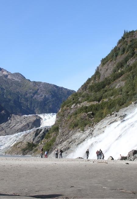Vor einer breiten Gletscherfront des Mendenhall Glacier stehen Besucher am grauen Sandstrand unter klarem Himmel.