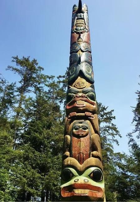 Ein hoher, bunt geschnitzter Totempfahl steht zwischen Nadelbäumen unter klarem Himmel im Sitka National Historical Park.