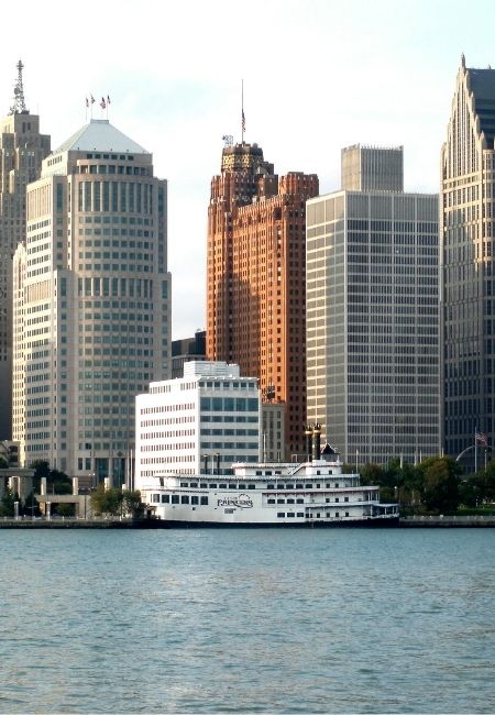 Die Skyline von Detroit erhebt sich am Wasser, während ein weißes Passagierschiff vor den Hochhäusern entlangfährt.