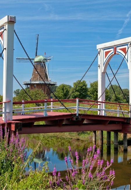 Eine rote Hängebrücke führt über einen Wasserlauf, dahinter steht eine Windmühle unter blauem Himmel in Holland, Michigan.