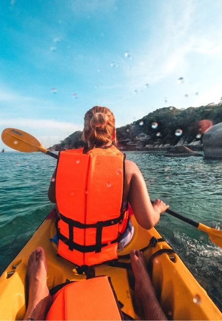 Eine Person paddelt im gelben Kajak mit orangefarbener Schwimmweste über ruhiges Wasser vor einer grünen Küste.