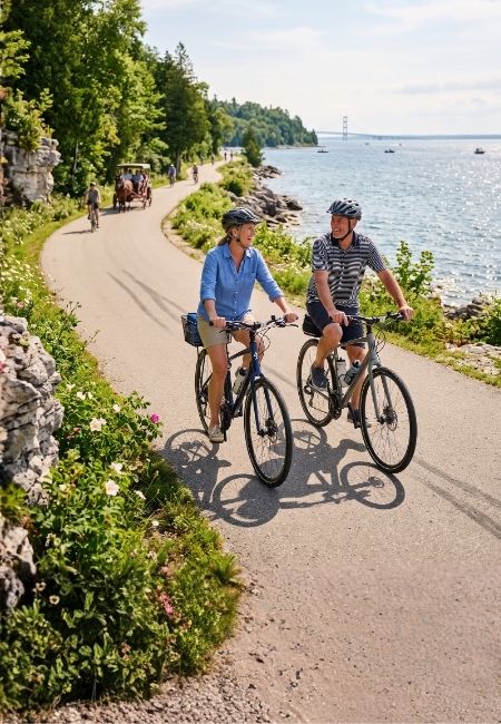 Zwei Radfahrer fahren auf Mackinac Island an einem kurvigen Uferweg entlang, mit See, Felsen und Grün im Hintergrund.