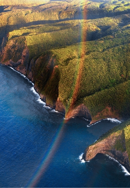 Grüne Küstenklippen fallen steil ins tiefblaue Meer, während ein Regenbogen über der zerklüfteten Landschaft verläuft.