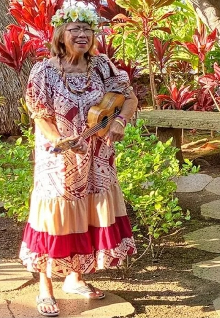 Eine lächelnde Frau mit Blumenkranz spielt Ukulele beim Hula-Luau zwischen roten und grünen Tropenpflanzen.