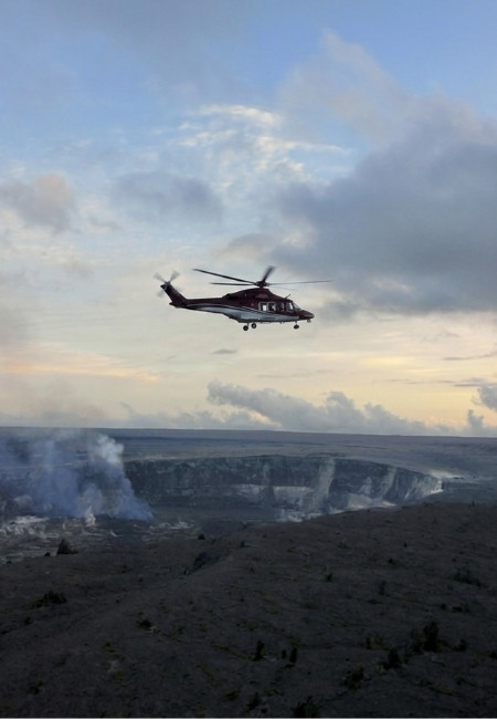 Über Big Island kreist ein Hubschrauber über einem rauchenden Vulkankrater und einer kargen Lavazone bei Sonnenlicht.