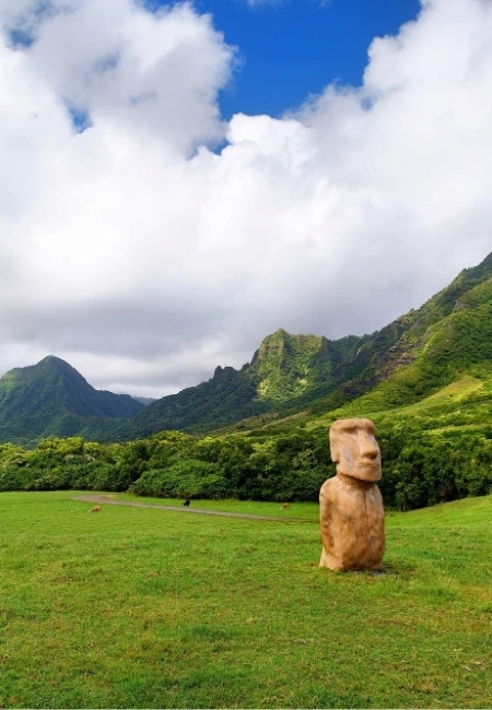 Auf der Kualoa Ranch auf Oʻahu steht eine Moai-Statue auf einer grünen Wiese vor steilen, üppig bewachsenen Bergen.