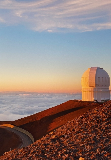 Am Mauna Kea auf Hawaii steht ein Observatorium über einem vulkanischen Hang, darunter liegt ein Wolkenmeer im Licht.