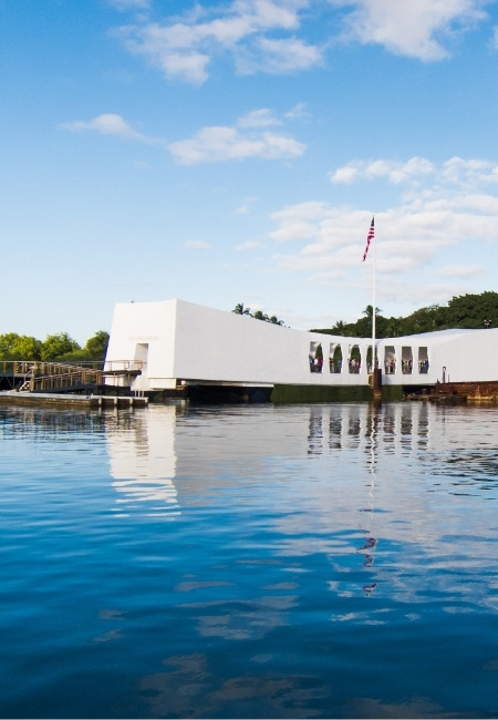 Das weiße USS Arizona Memorial in Pearl Harbor spiegelt sich im ruhigen Wasser, daneben weht die US-Flagge.