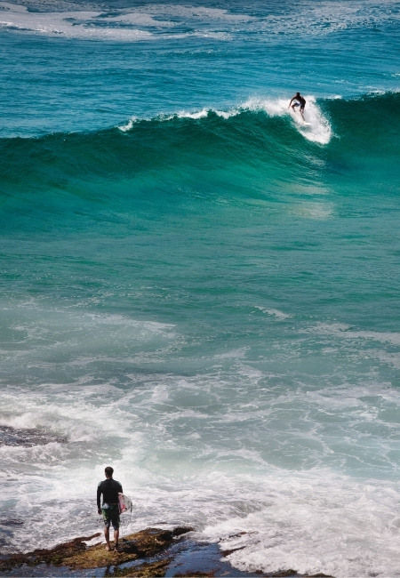 An der North Shore von Hawaii reitet ein Surfer eine kräftige Welle, während am Ufer ein weiterer mit dem Brett wartet.