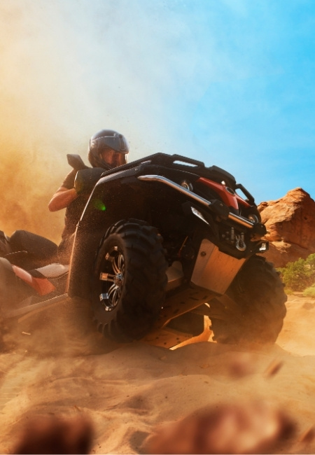 Ein Fahrer lenkt ein ATV durch sandiges Gelände in der Mojave, während eine große Staubwolke und rote Felsen die Szene prägen.
