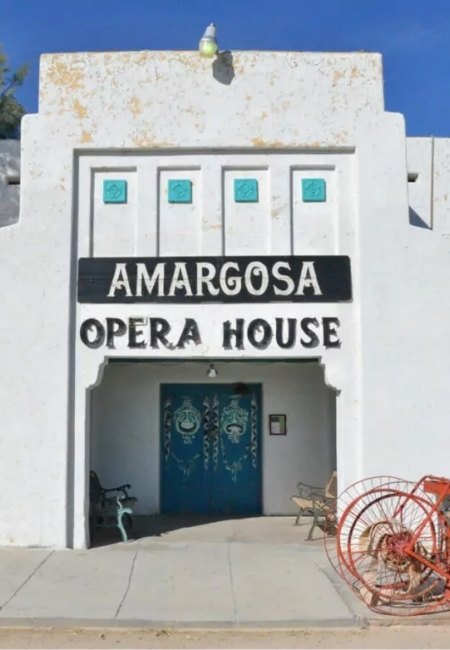 Vor der weißen Fassade des Amargosa Opera House stehen ein blaues Doppeltor, kleine Sitzbänke und ein rostiges Fahrrad im hellen Wüstensonnenschein.
