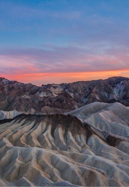 Die zerklüfteten Fels- und Sandformationen im Death Valley leuchten unter einem rosa Abendhimmel, während die Berge im Hintergrund dunkel wirken.