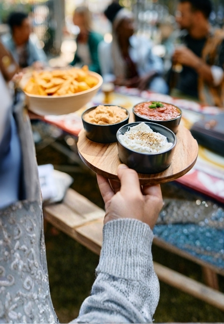 Eine Hand reicht auf einer Las Vegas Food-Tour ein Holztablett mit drei Dips und Pommes, während im Hintergrund Gäste stehen.