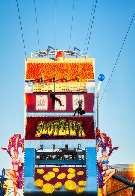 Vor der leuchtenden SlotZilla-Fassade an der Fremont Street in Las Vegas schweben zwei Zipline-Fahrer unter Kabeln vor blauem Himmel.