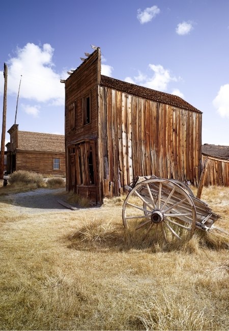 In der Geisterstadt Bodie steht ein verwittertes Holzhaus neben einem alten Wagenrad im trockenen Gras unter blauem Himmel.